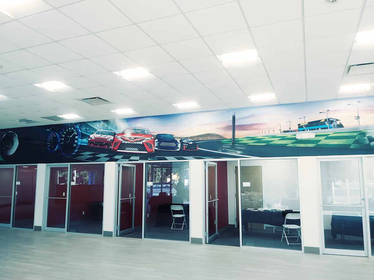 Modern office space with glass-walled rooms and a large mural of racing cars and a race track above.