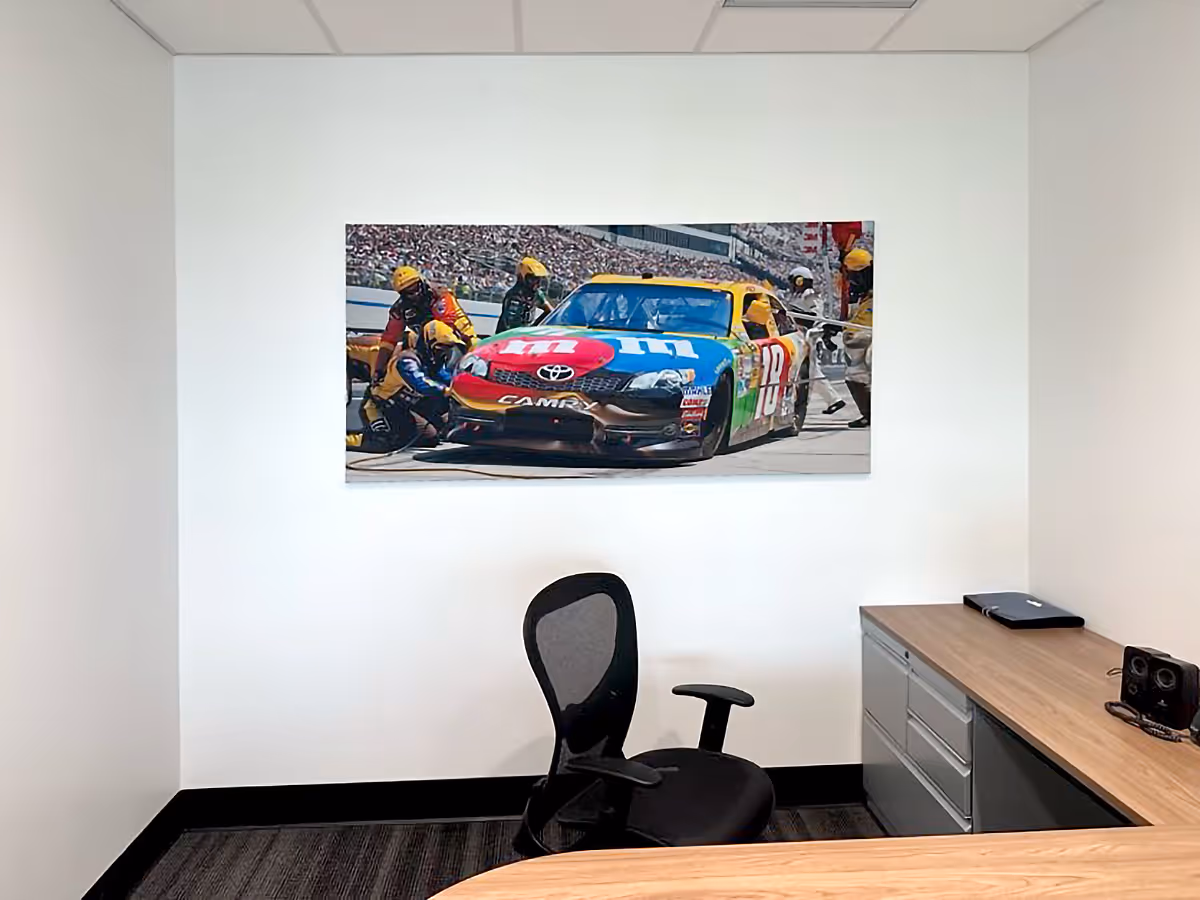 Small office with black mesh chair, wooden desks, and a Silicone Edged Graphic Frame with a colorful fabric print of M&M race car poster on a white wall.