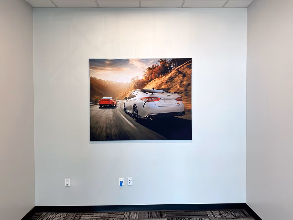 Silicon Edged Graphic Framed Fabric print of a white Toyota Camry and a red car driving on a winding road during sunset, mounted on a plain office wall.