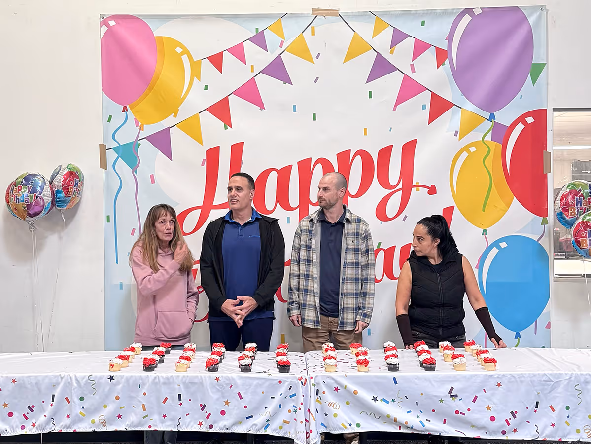 Four people standing behind a table with cupcakes in front of a colorful 'Happy Birthday' banner with balloons and bunting decorations.