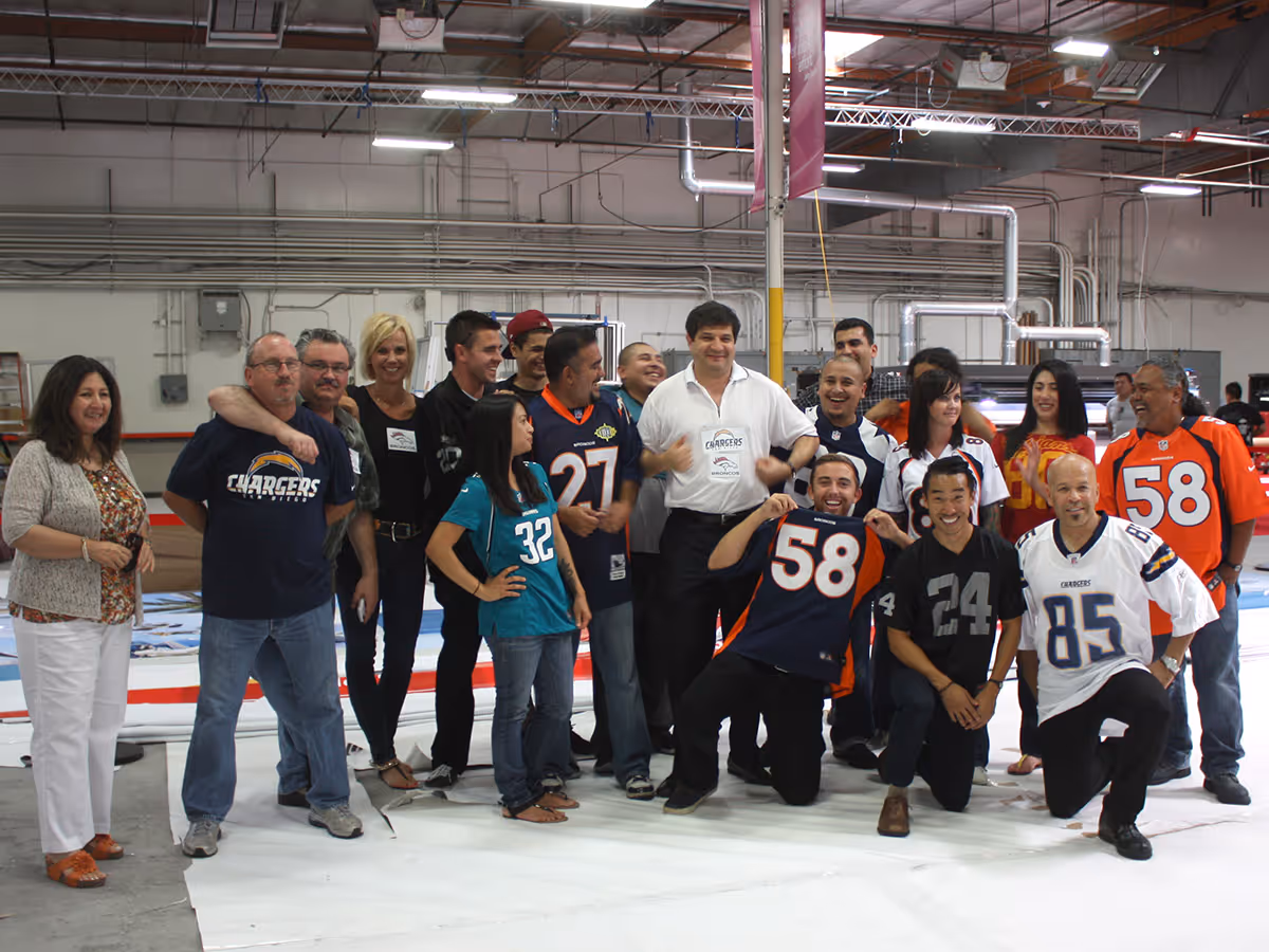 Group of people wearing various football jerseys posing together indoors in a warehouse-like setting.