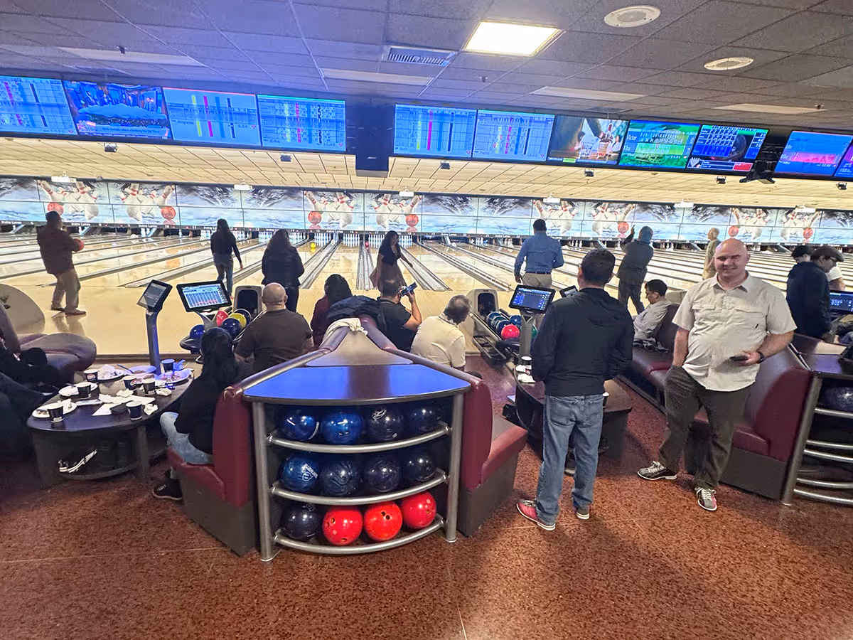 People socializing and bowling in a busy bowling alley with multiple lanes and screens overhead.