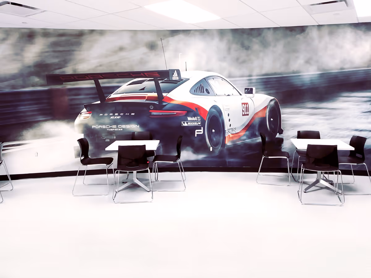 Modern white room with black chairs and tables in front of a large mural of a Porsche 911 race car drifting with smoke.