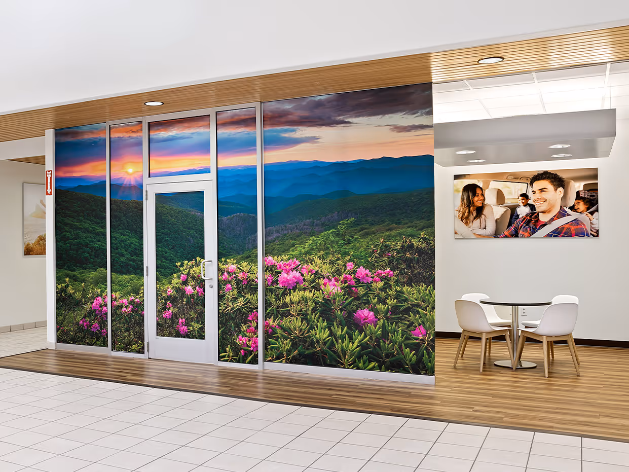 Indoor space with wall-sized window graphics showing a mountain landscape with pink flowers at sunset and a seating area with a photo of a smiling family inside a car.