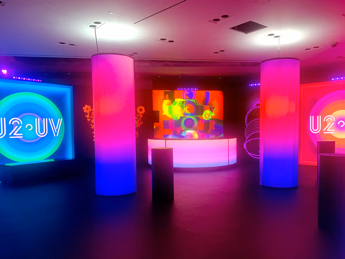 Colorful illuminated exhibition space with cylindrical light pillars and neon U2 UV signs on the walls.