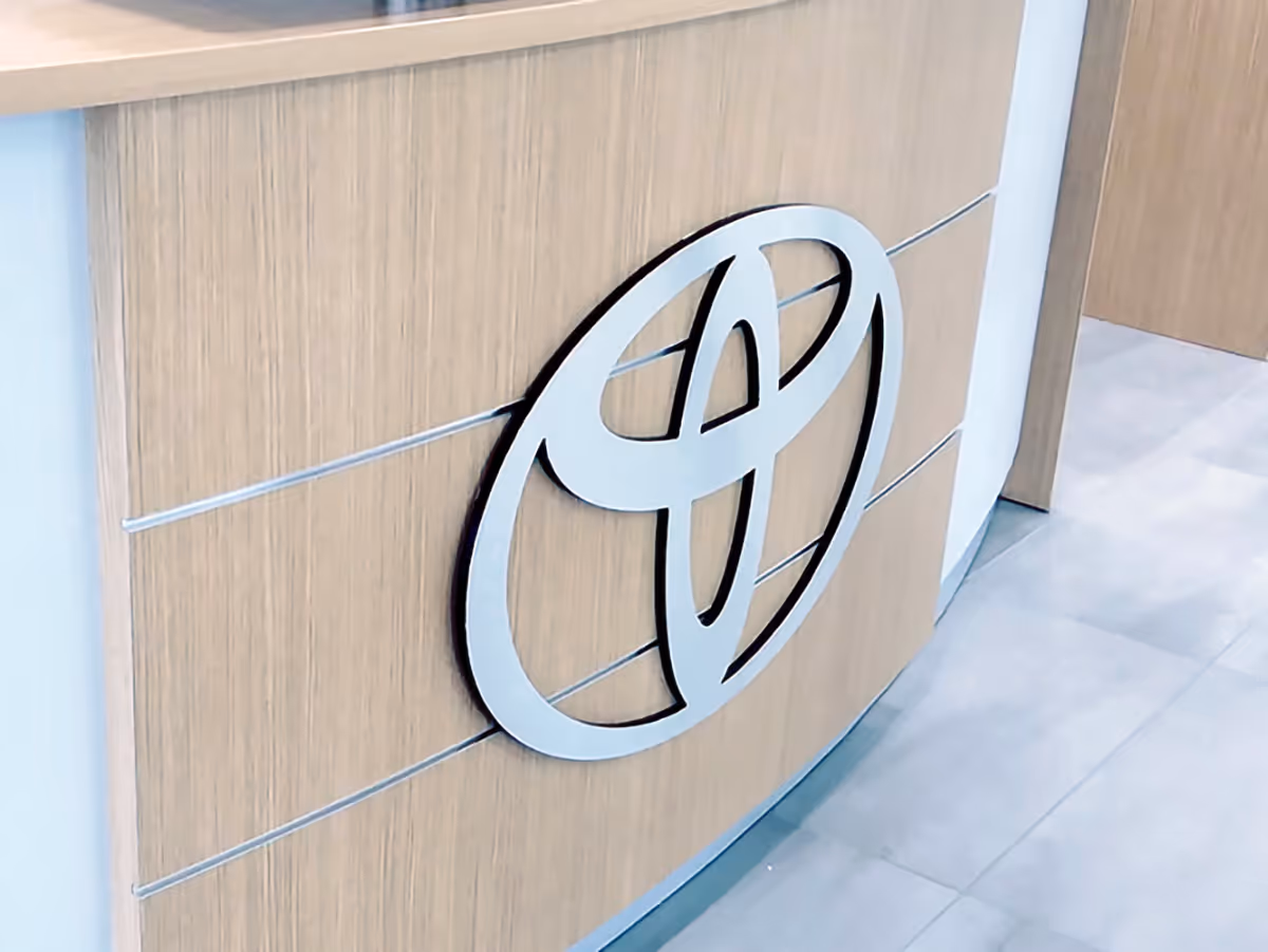 Silver Toyota fabricated logo mounted on a light wooden curved wall panel with gray tiled floor.