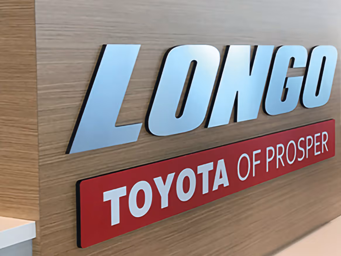 Fabricated Sign on a wooden wall reading 'LONGO' in large silver letters above a red plaque with 'TOYOTA OF PROSPER' in white letters.