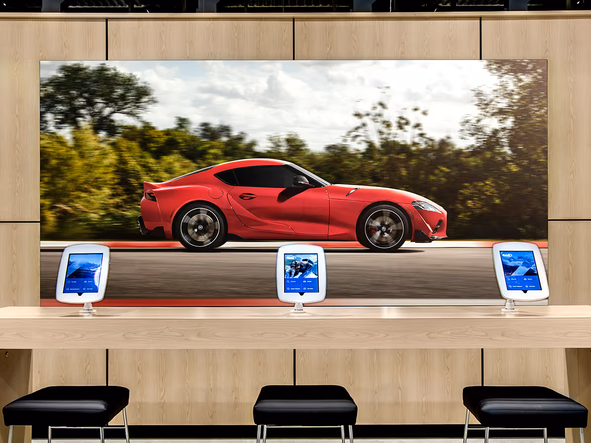 A large LED backlit print of Red sports car driving past trees displayed above a wooden counter with three tablets and black stools.