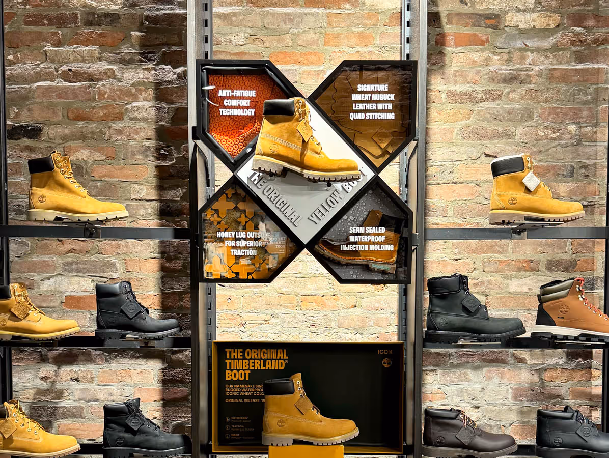 Display of Timberland boots including the classic yellow boot with features like anti-fatigue comfort and waterproof injection molding highlighted on a brick wall background.