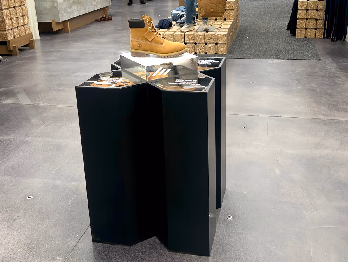 Yellow Timberland-style boot displayed on a black hexagonal pedestal in a store with wooden crates and carpeted area in the background.