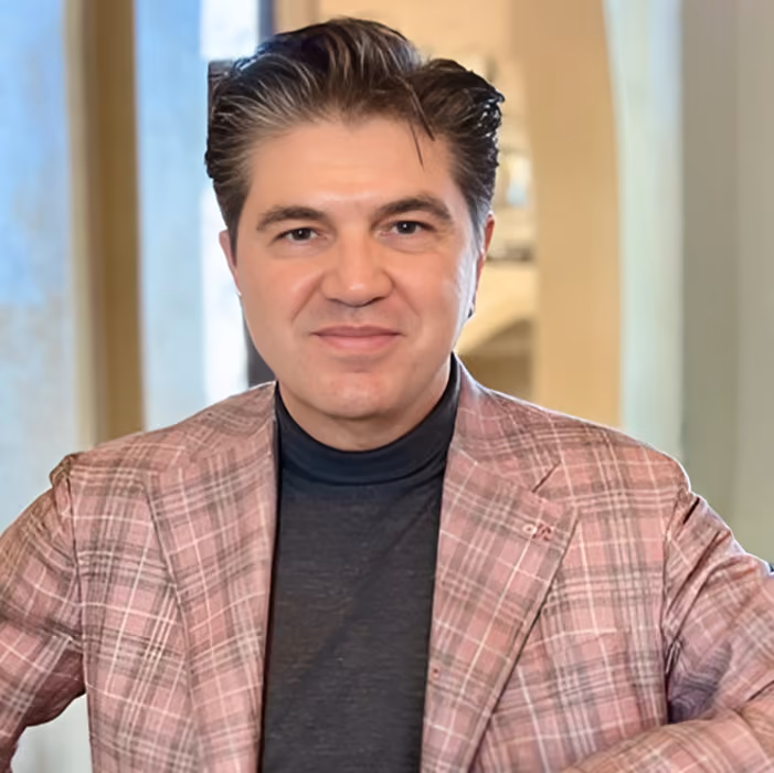 Man in a pink plaid blazer and black turtleneck holding a glass of white wine and smiling.