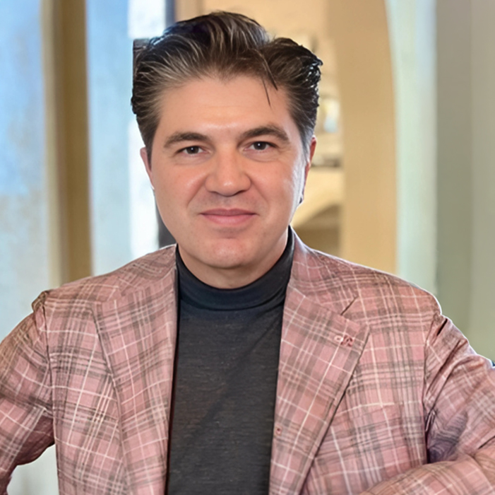 Man in a pink plaid blazer and black turtleneck holding a glass of white wine and smiling.