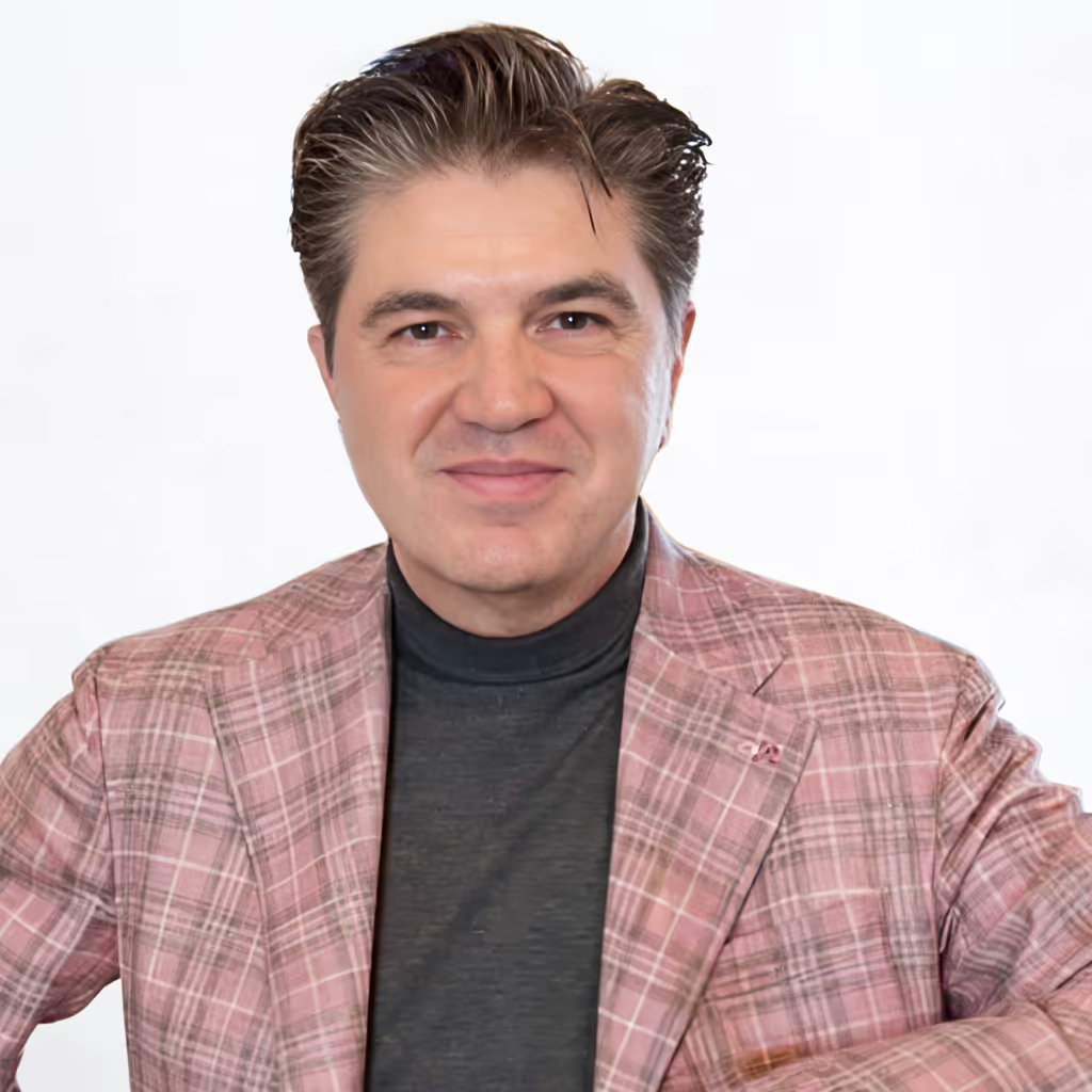 Man in a pink plaid blazer and black turtleneck holding a glass of white wine and smiling.