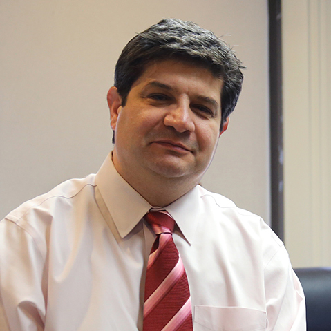Man with dark hair wearing a white shirt and striped red tie sitting indoors in front of a window.