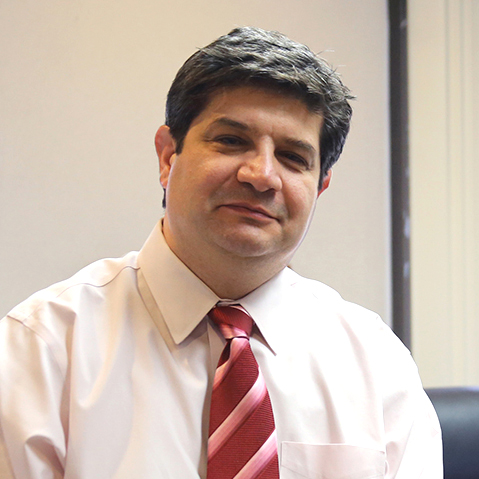 Man with dark hair wearing a white shirt and striped red tie sitting indoors in front of a window.