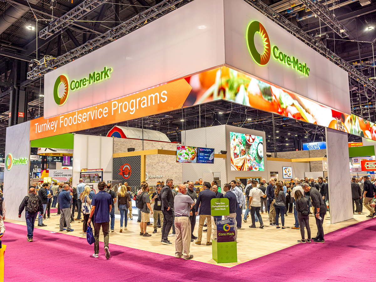 Busy Core-Mark trade show booth with many people walking and mingling on a purple carpeted floor.