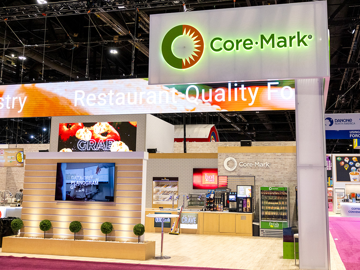 Trade show booth for Core-Mark featuring a large illuminated logo, digital displays, and a refreshment area.