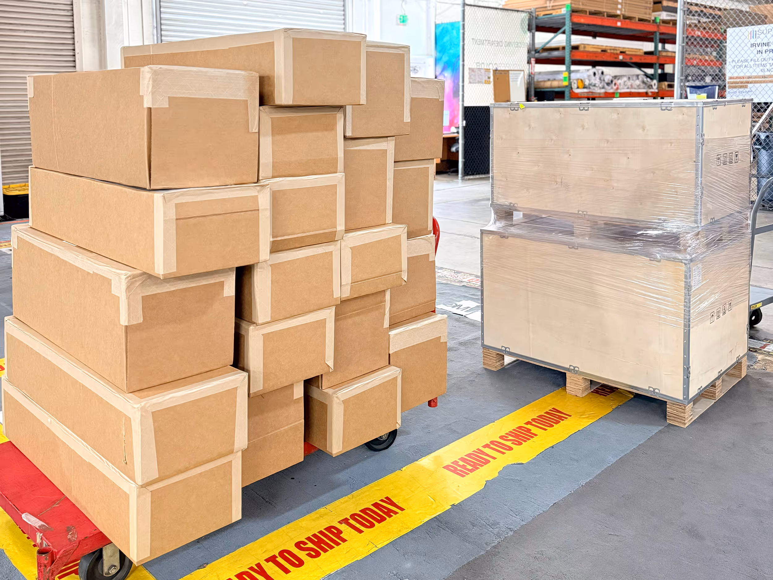 Stack of taped cardboard boxes and wrapped wooden crates on pallets in a warehouse near a yellow floor marking that says 'READY TO SHIP TODAY'.