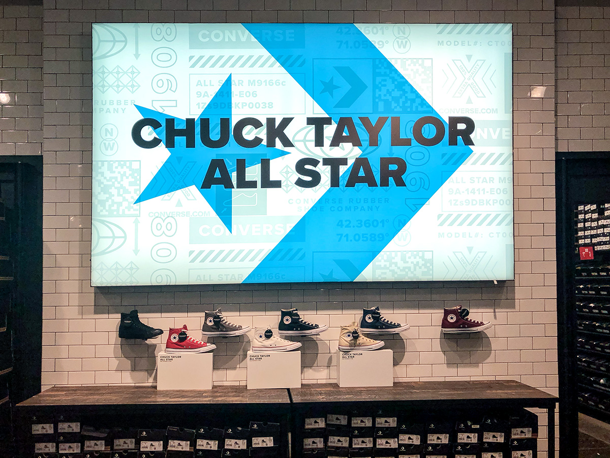 Yellow Timberland-style boot displayed on a black hexagonal pedestal in a store with wooden crates and carpeted area in the background.
