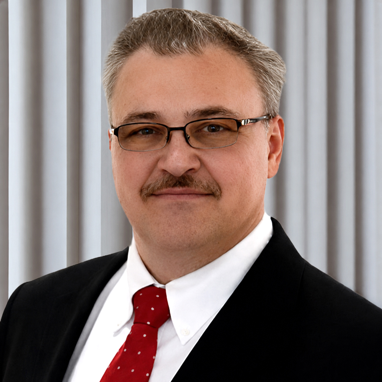 Man wearing glasses, a white shirt, red tie, and dark suit jacket standing in front of a colorful background with red and yellow hues.