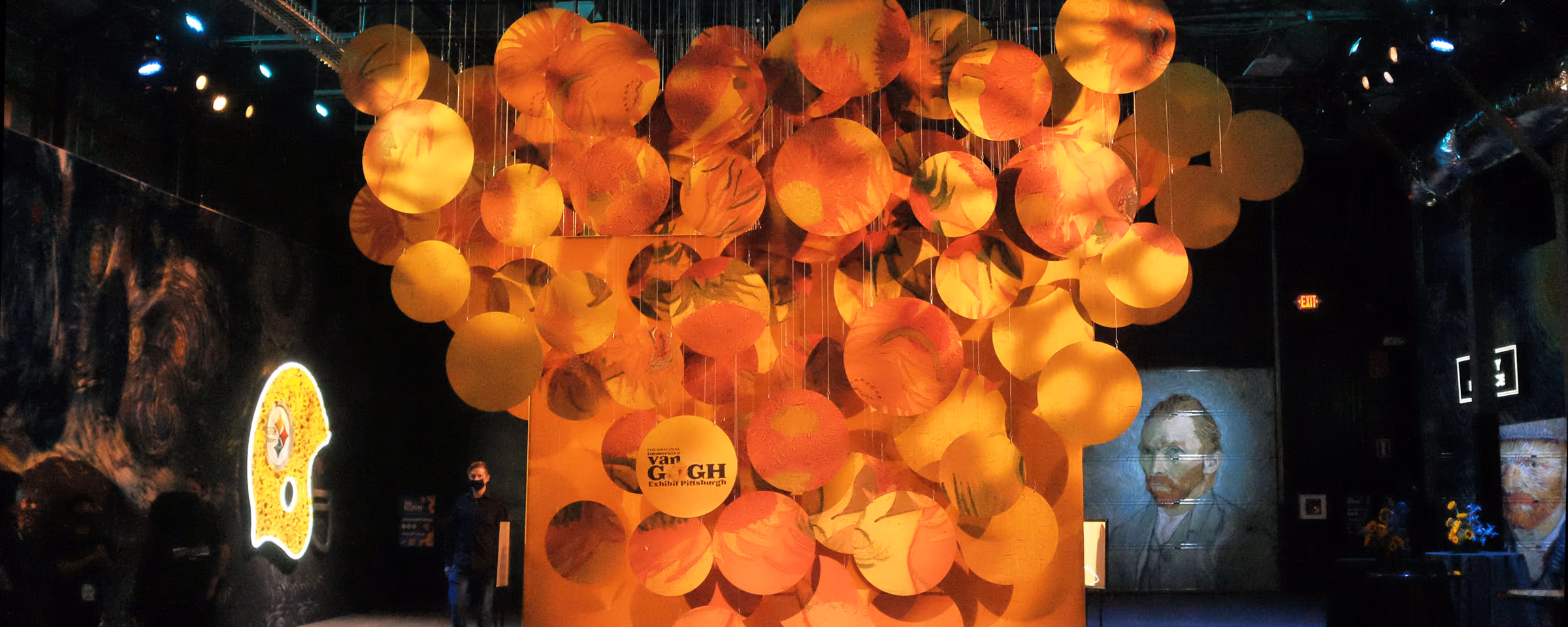 Indoor exhibit with orange and yellow circular discs hanging from the ceiling, illuminated against a dark background, featuring a sign that says 'The Original Van Gogh Exhibit Pittsburgh' and a projection of a Van Gogh self-portrait on the wall.