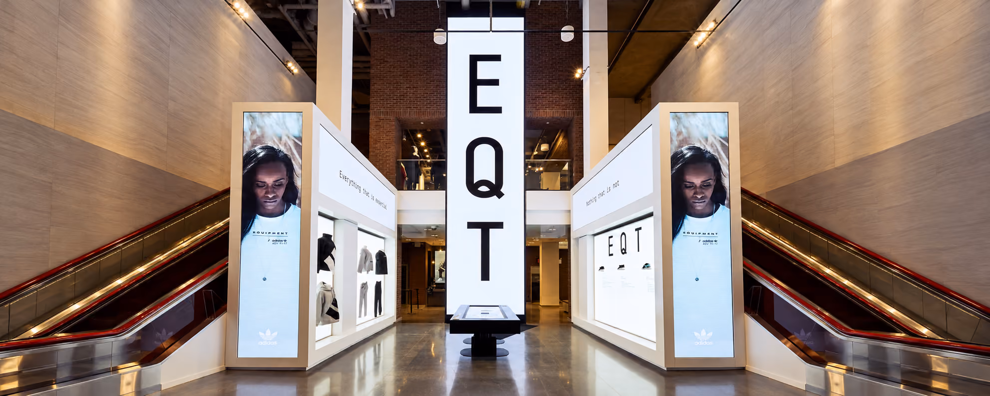 Modern retail space with two escalators on each side and a central display featuring large vertical signage with the letters EQT and digital SEG Lightbox panels showing a woman wearing an Adidas Equipment shirt, along with shelving displaying clothing items and shoes under soft lighting.