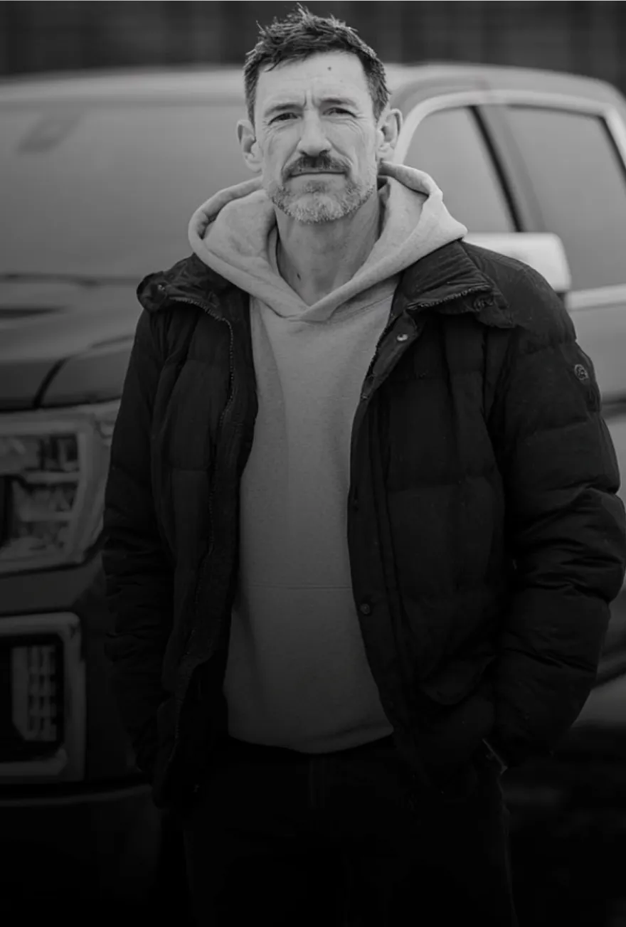 Middle-aged man with short hair and a beard wearing a hoodie and a puffer jacket standing in front of a pickup truck.