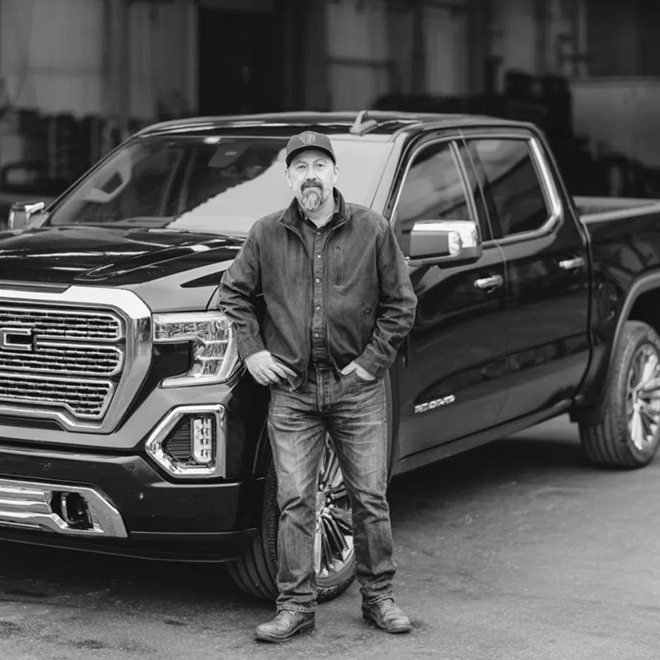 Man standing with hands in pockets in front of a black pickup truck inside a garage.