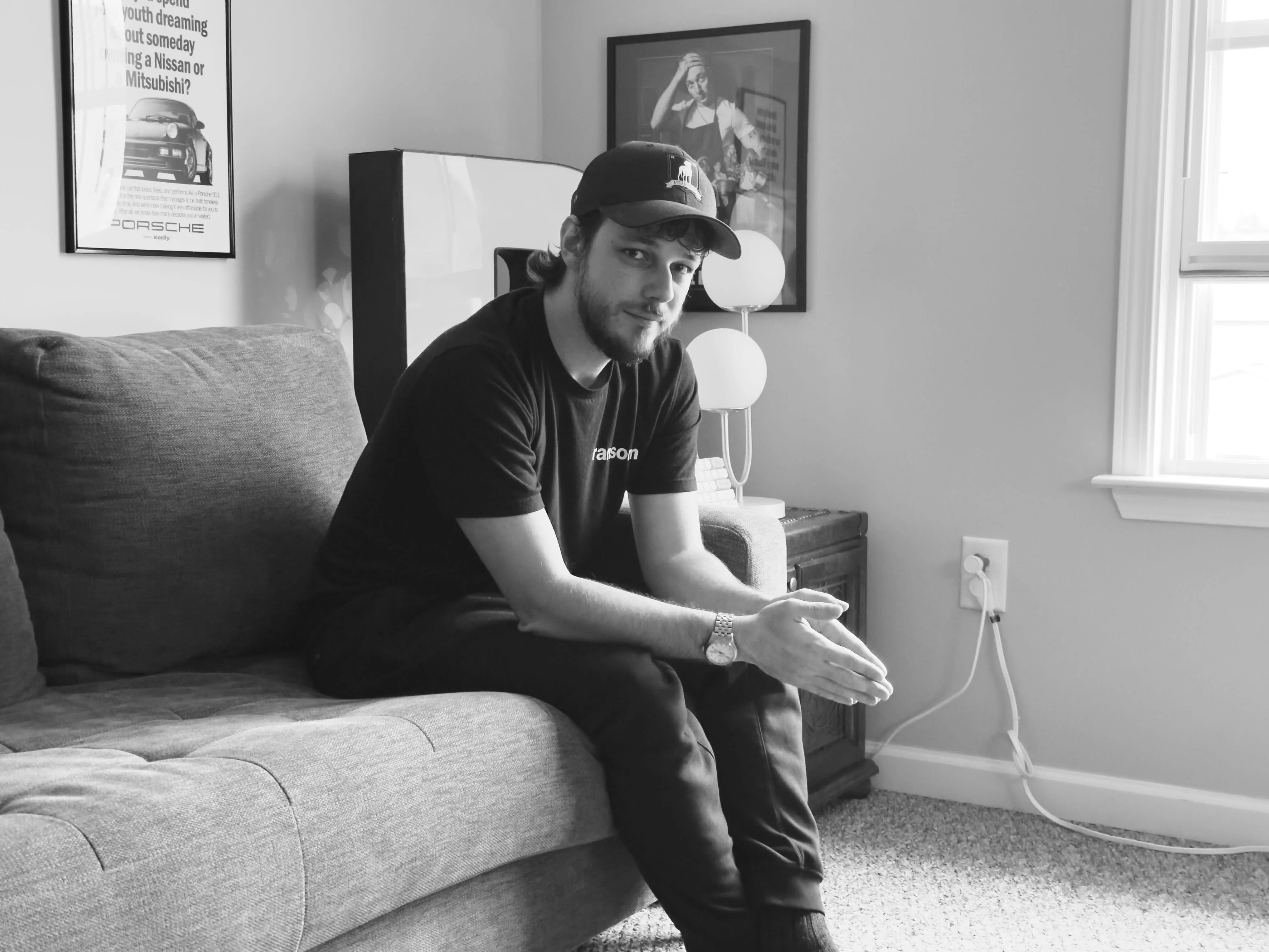 Young man wearing a cap and black clothes sitting on a couch with hands clasped, looking at the camera in a living room.