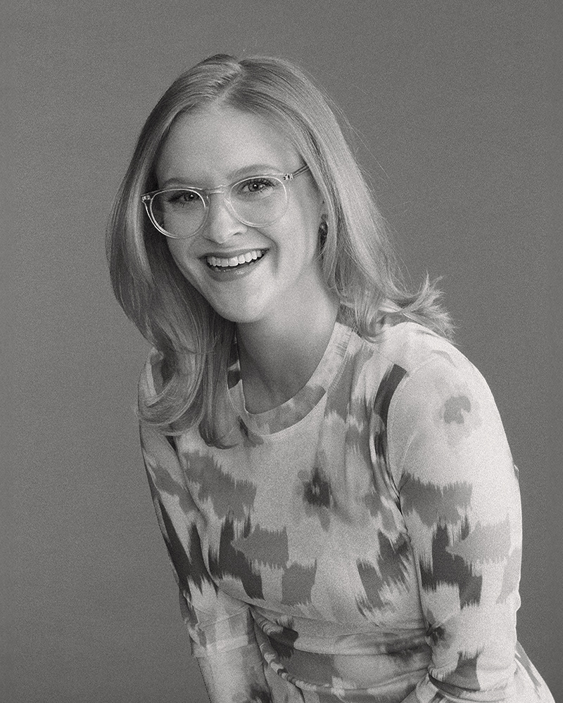 Smiling woman with shoulder-length hair and glasses wearing a patterned long-sleeve top.