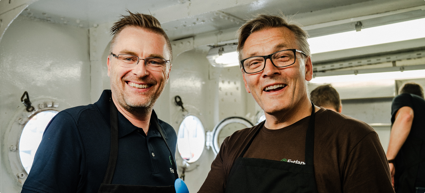 To menn med briller smiler og ser mot kameraet, iført svarte forkle i et lyst rom med runde vinduer.