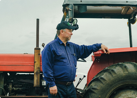 Mann i blå skjorte og svart caps som lener seg på en rød traktor ute under skyet himmel.