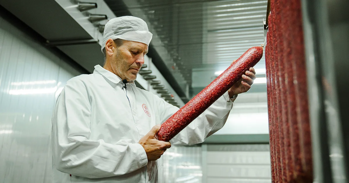 En mann i hvit frakk og hette inspiserer en stor spekepølse som henger i et kjølerom.