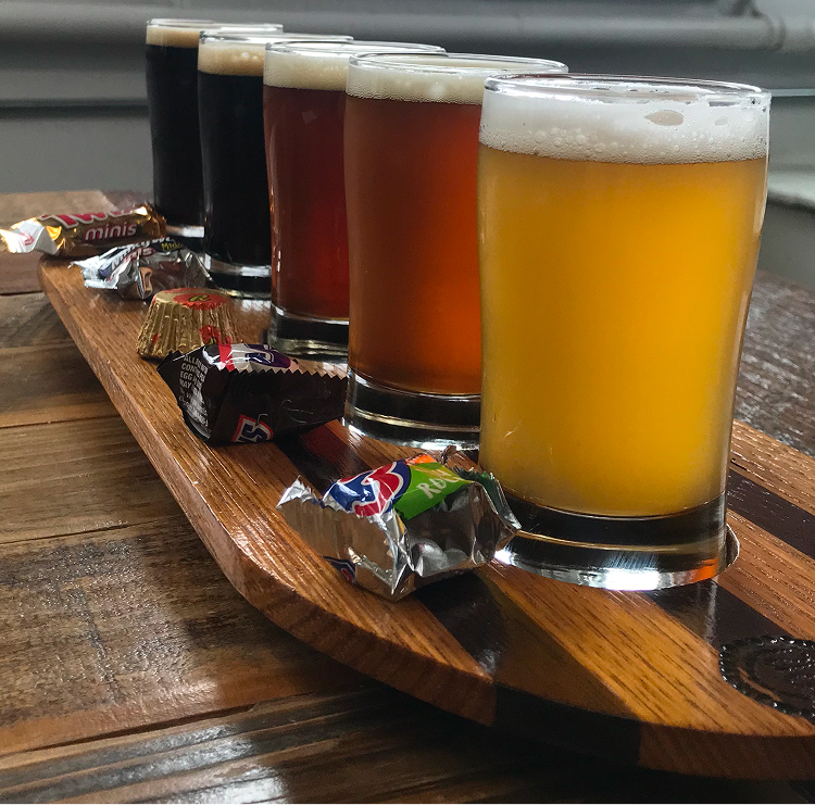 Flight of five beers in varying colors from dark to pale amber, served on a wooden paddle with candies.