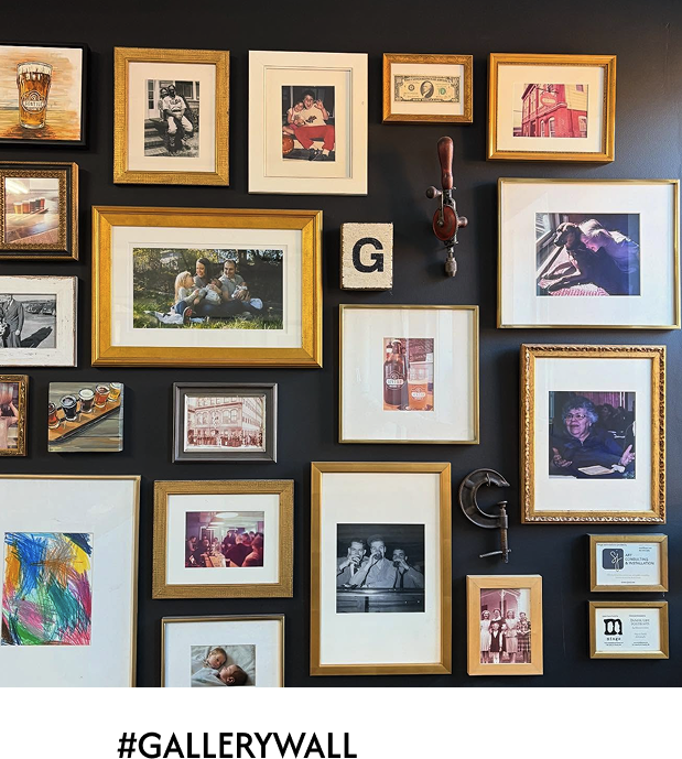 Gallery wall with framed photos, artwork, a dollar bill, and vintage tools on black background.