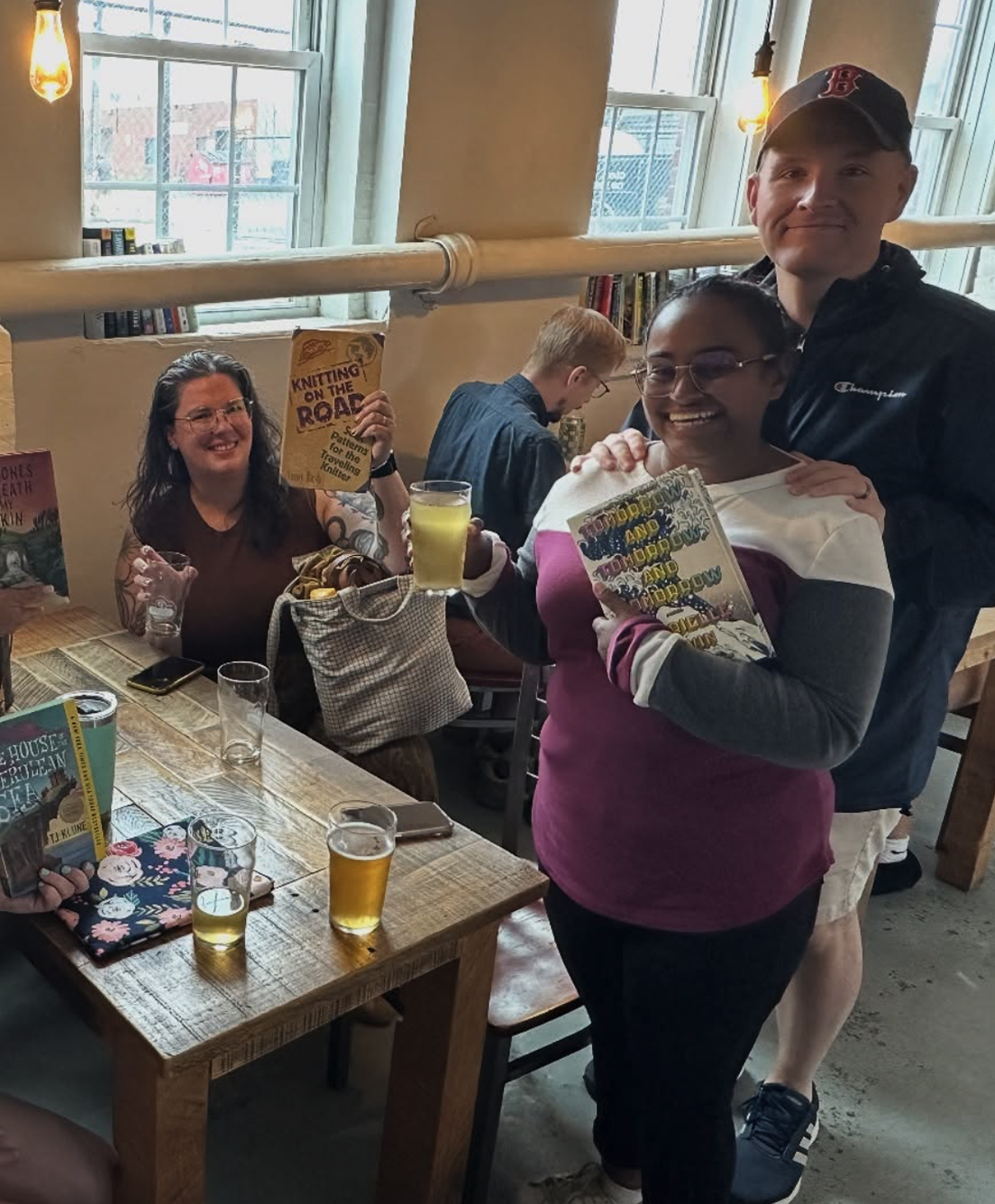 Four people holding books and drinks at a wooden table in a cozy, sunlit room.