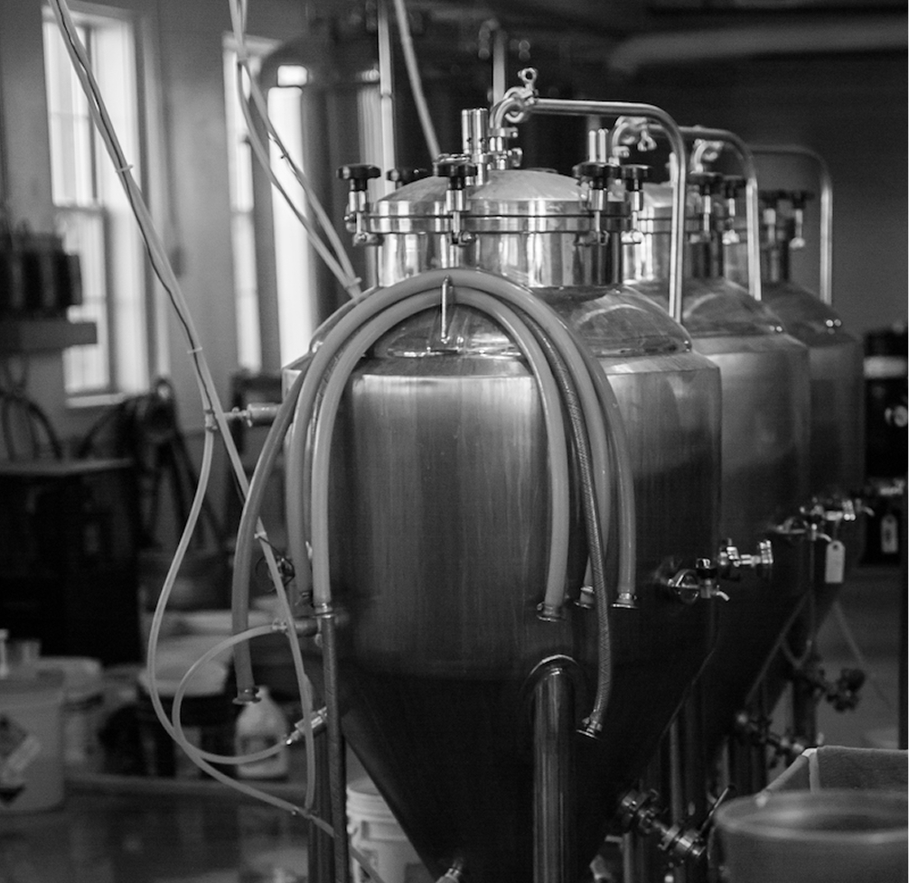 Row of stainless steel fermentation tanks with hoses in a brewery or industrial setting.