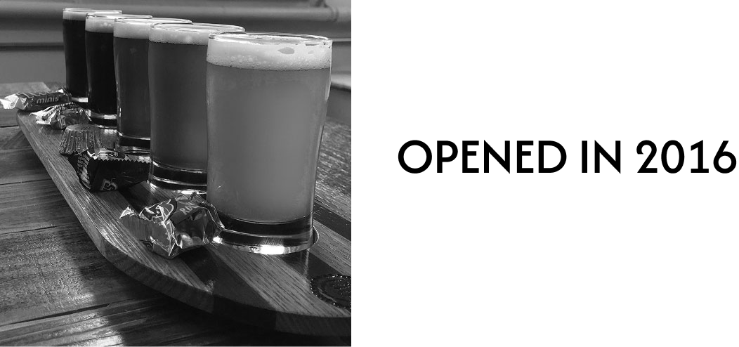 Six beer glasses with foam on a wooden paddle, each paired with wrapped chocolates.