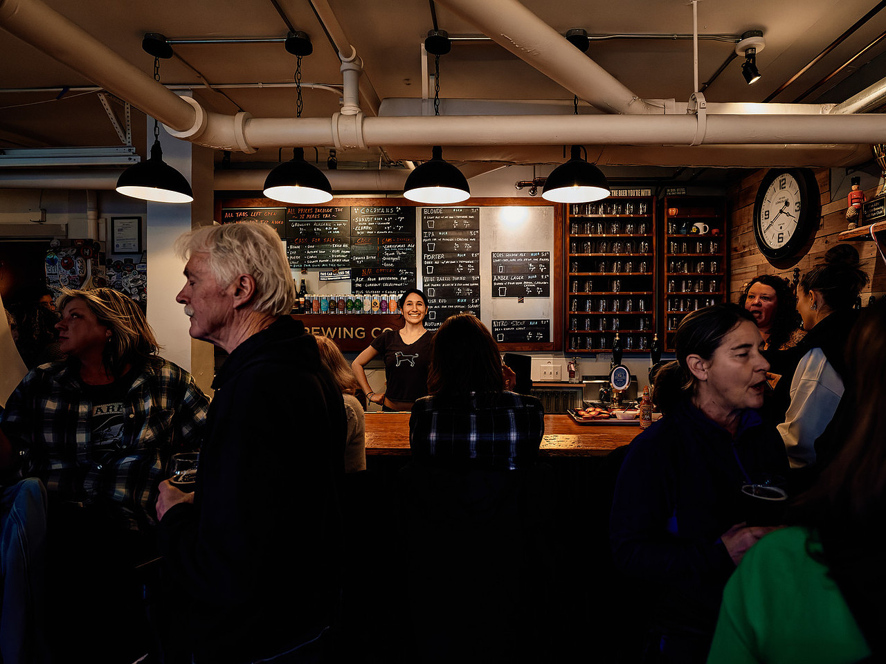 People socializing at a bar with a smiling bartender and a menu board in the background.