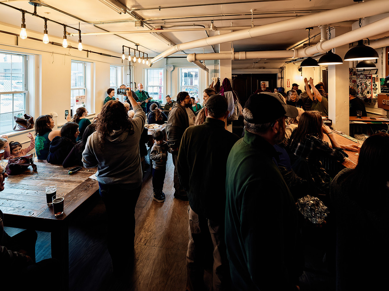 Crowded brewery with people watching a live music performance and socializing indoors.