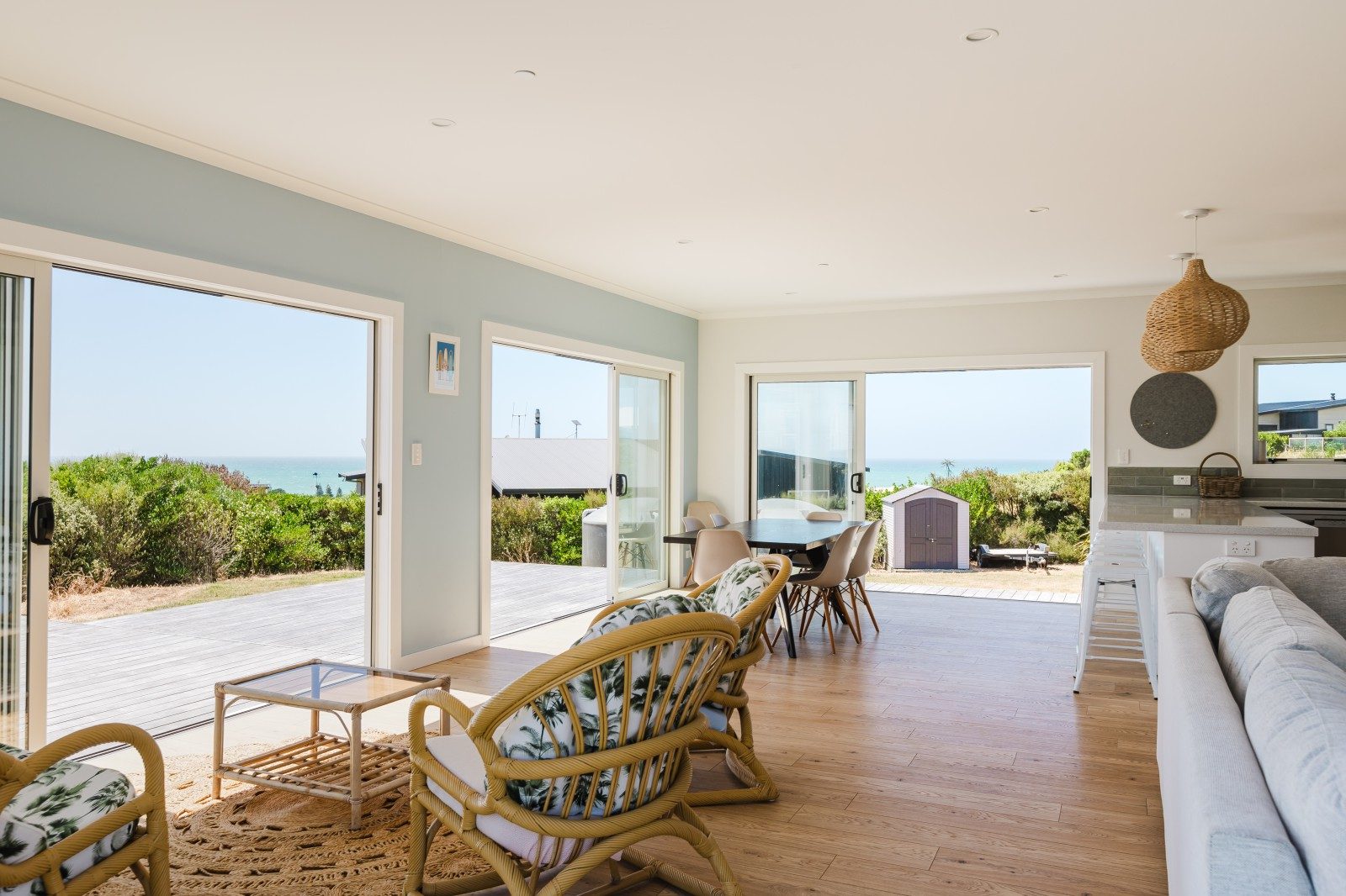 Bright living and dining area with rattan chairs, wooden floors, large sliding glass doors opening to a deck and ocean view.