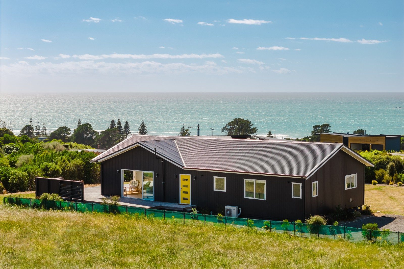 Modern single-story dark gray house with a yellow door overlooking the ocean and surrounded by green shrubbery and grass.
