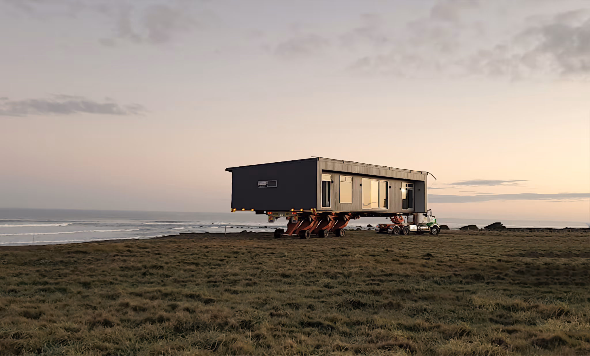 Large modern house elevated on hydraulic lifts attached to a truck, set on grassy land near the ocean at sunset.