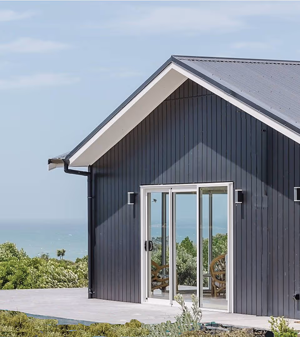 Modern dark gray wooden house with large sliding glass doors overlooking a green landscape and ocean.