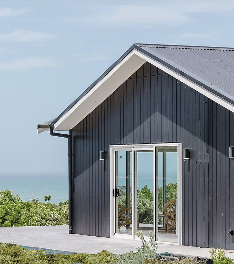 Modern dark gray wooden house with large sliding glass doors overlooking a green landscape and ocean.