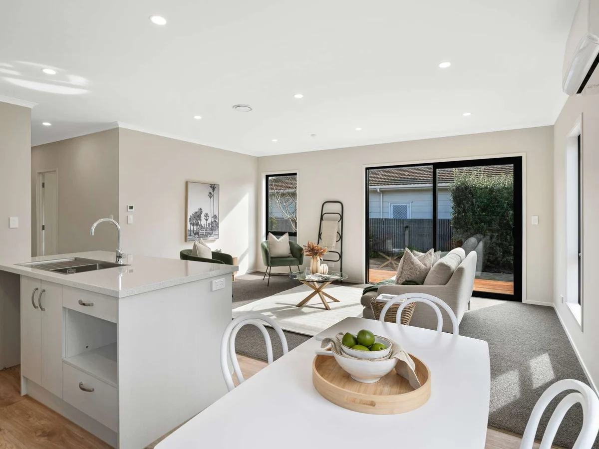 Modern open-plan living area with a white kitchen island, dining table with green apples centerpiece, and seating area with sofa and armchairs near large sliding glass doors.
