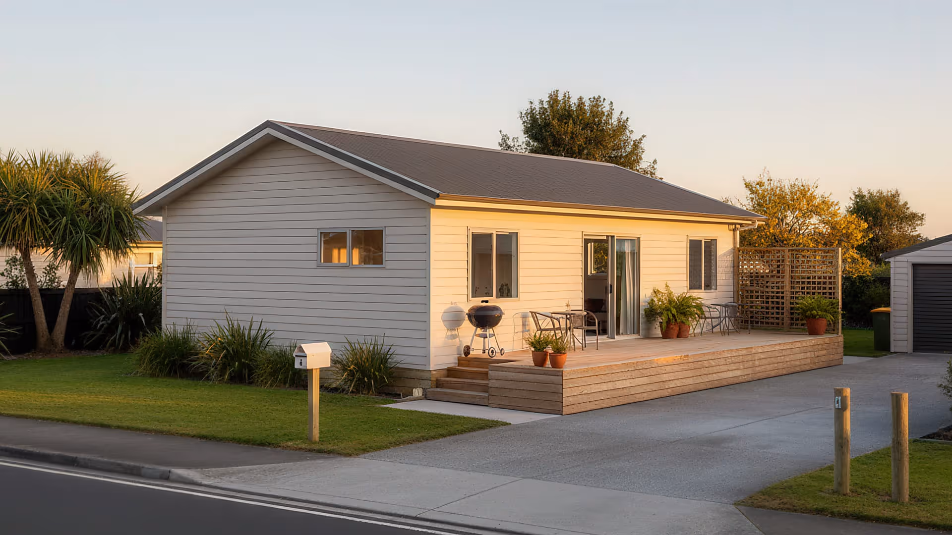 Single-story house with beige siding, a small wooden deck with potted plants, outdoor chairs, and grills, surrounded by a green lawn at sunset.