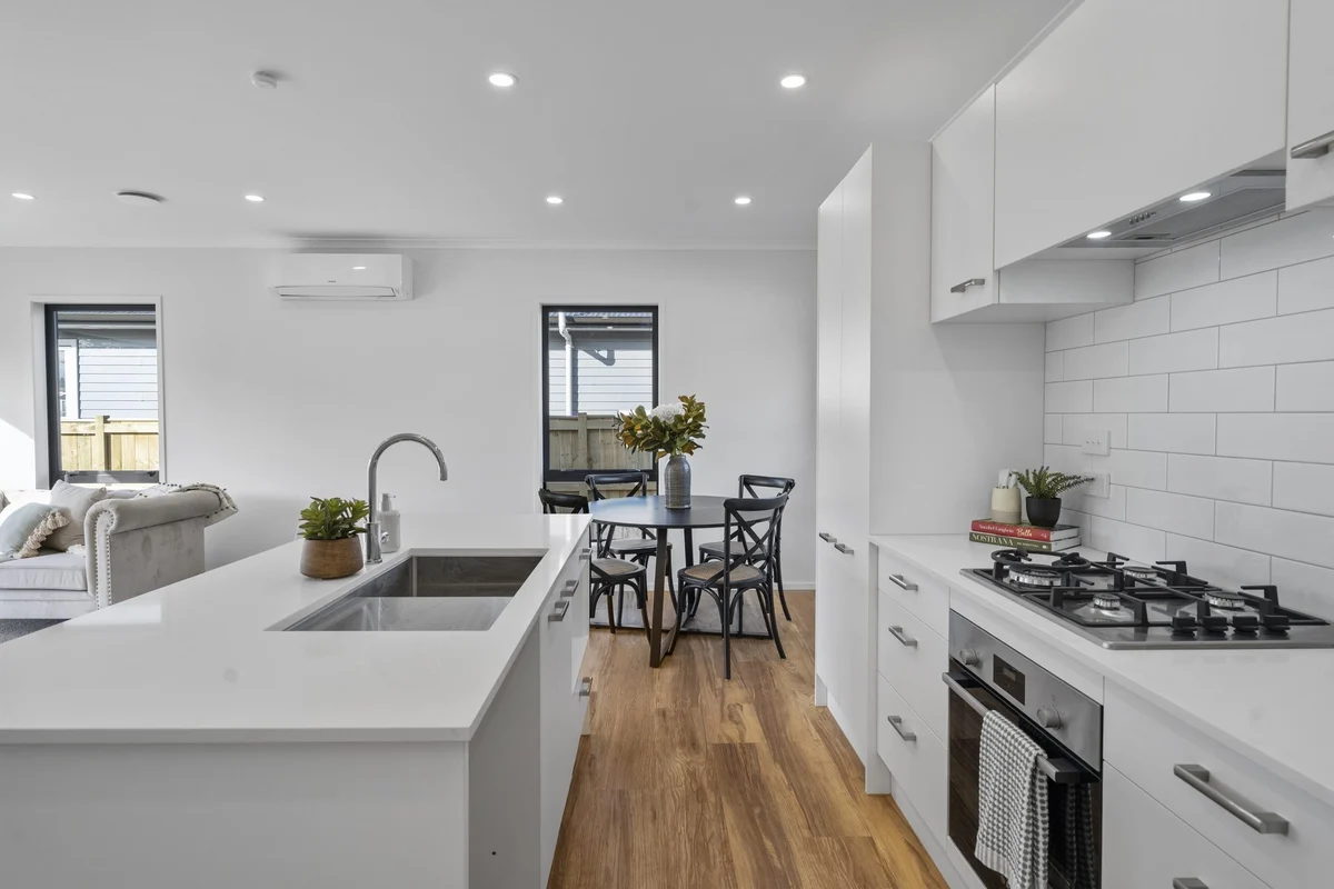 Modern kitchen with white cabinetry, a white island with a sink and plant, wooden floors, and a small dining table with black chairs and a vase of flowers.