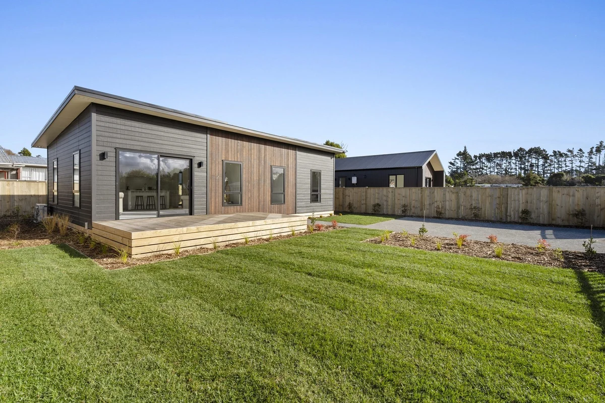 Modern single-story house with mixed wood and gray siding, a wooden deck, and a large green lawn under a clear blue sky.