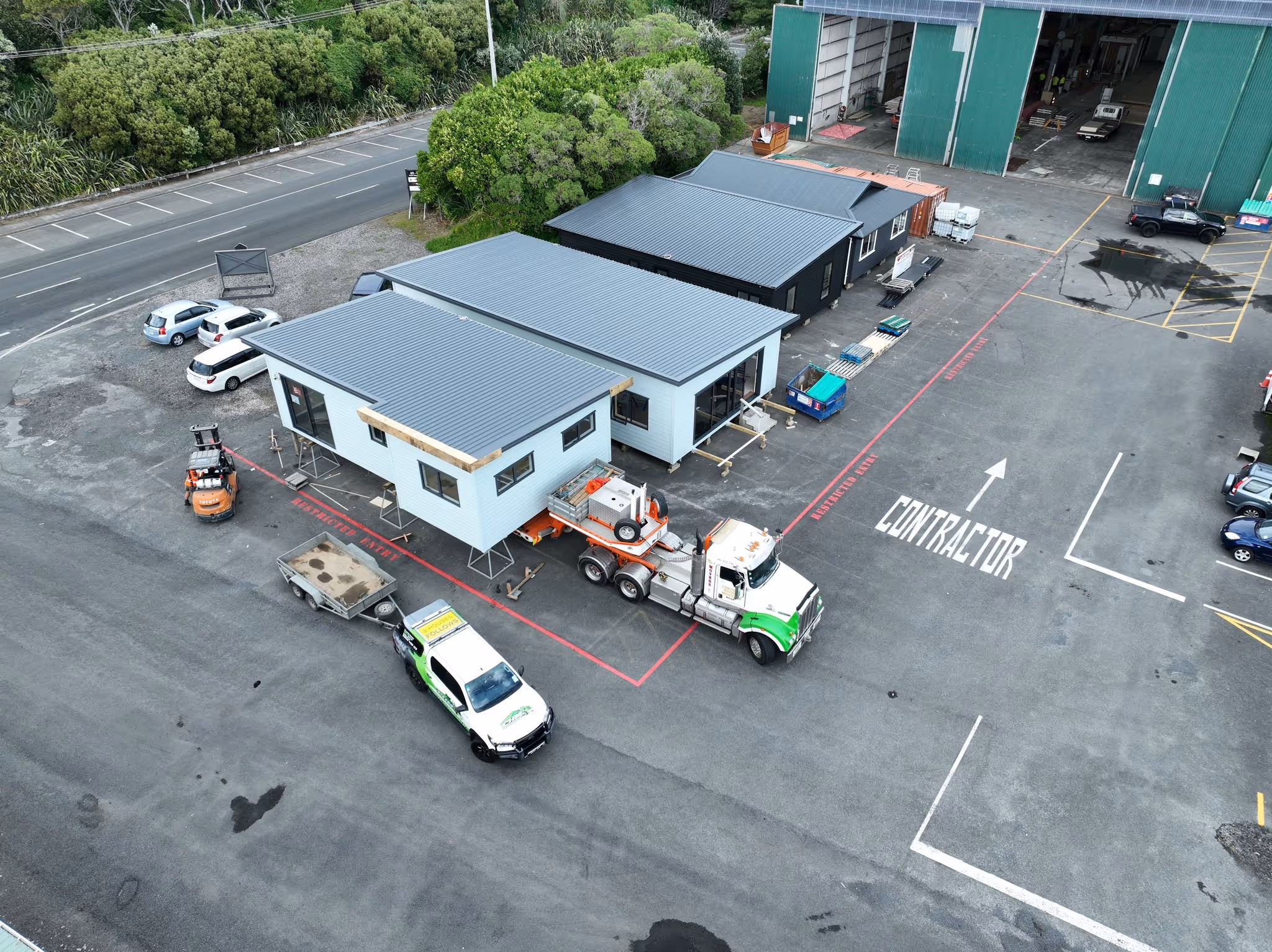 Aerial view of a factory yard with two prefabricated modular buildings on trailers, a white and green truck, forklift, several parked cars, and a large green warehouse in the background.
